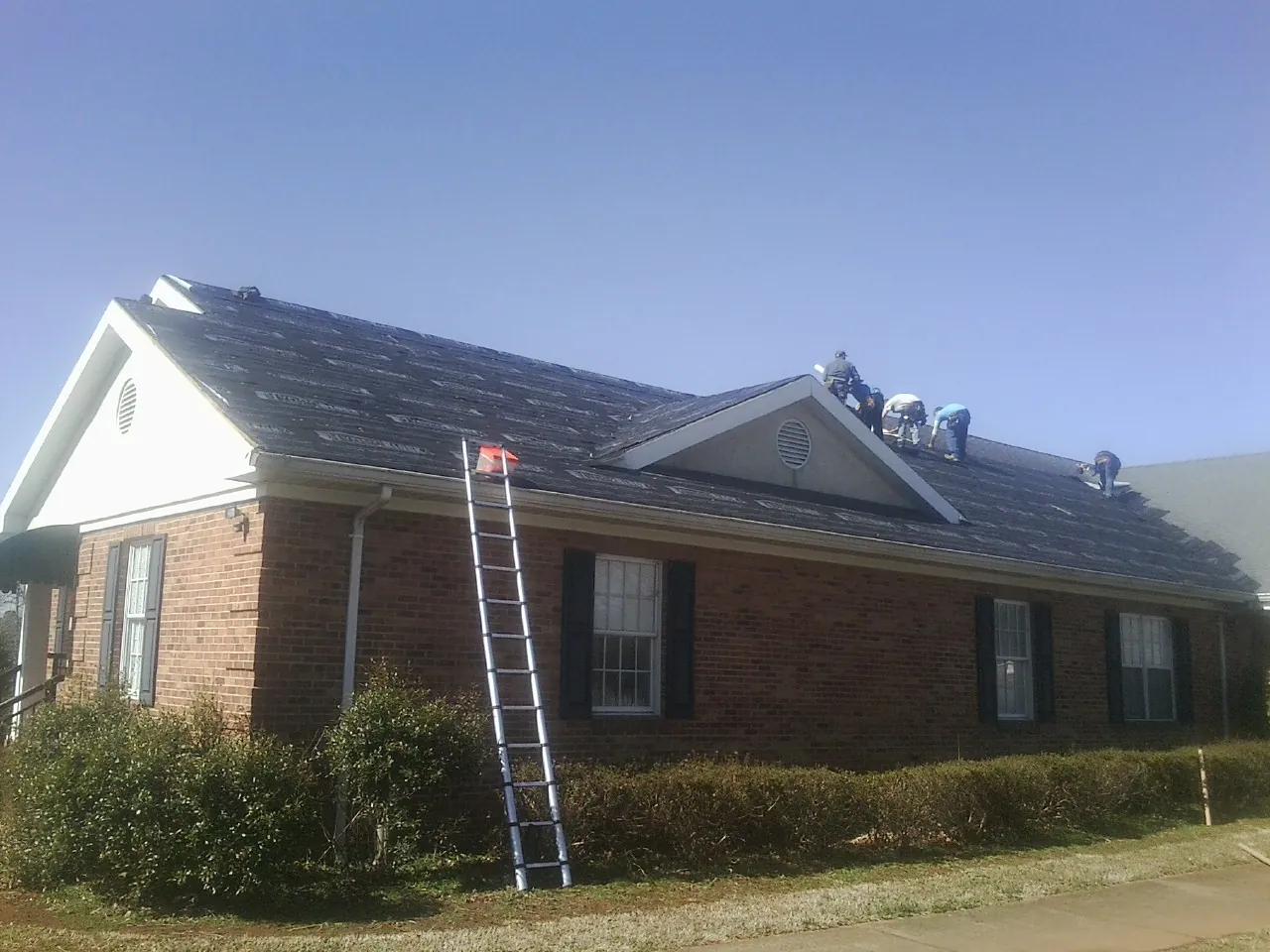Crew tearing shingles off a brick ranch roof with a ladder set — Emergency Roof Repair work by our crew in Rayne