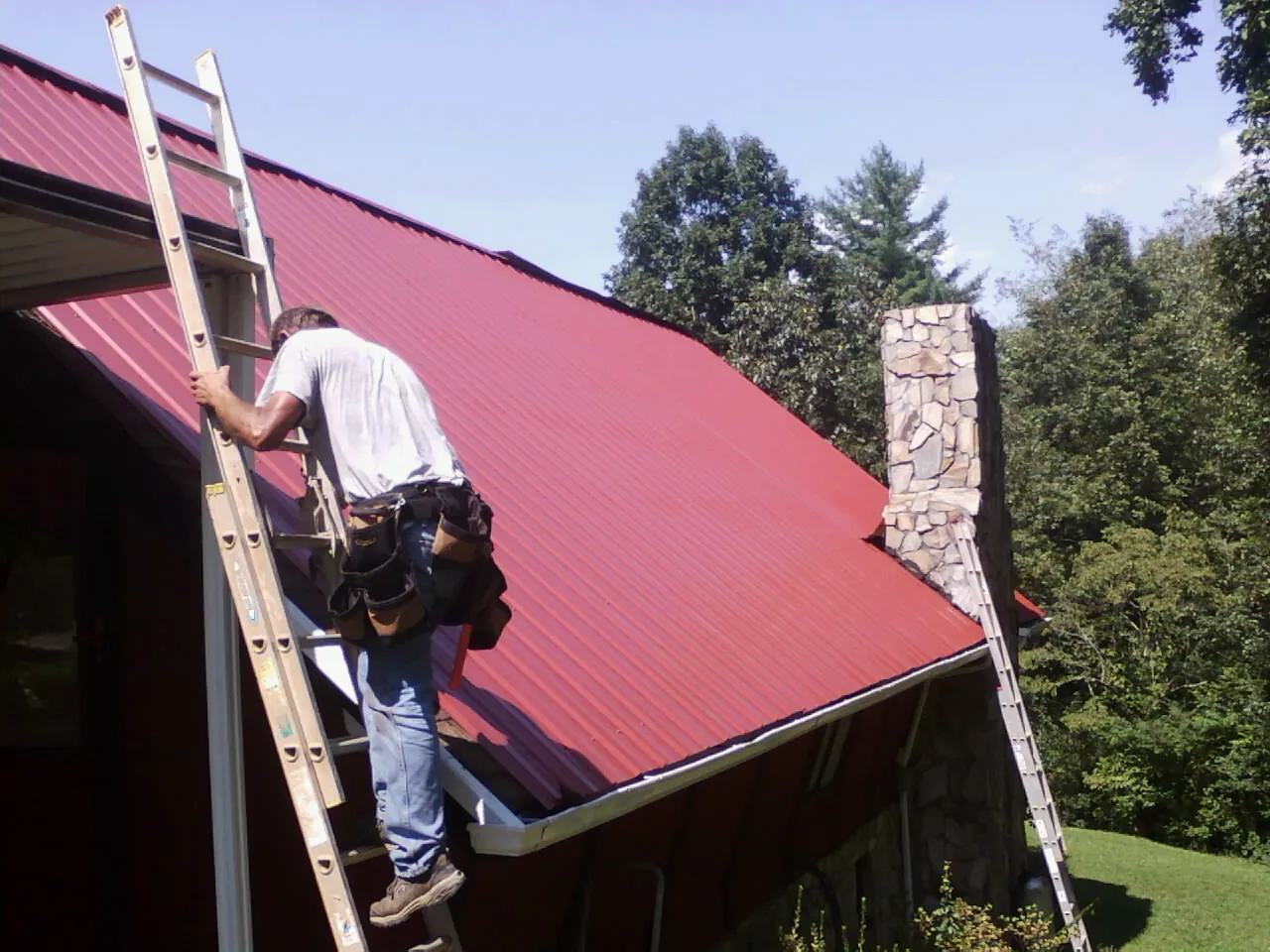 Roofer on a ladder beside a red metal roof and stone chimney — Soffit & Fascia Repair by Roofing Brothers in Rayne